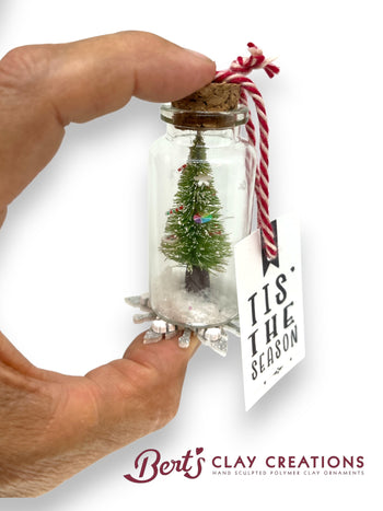 Hand holding a small glass jar with a miniature Christmas tree and 'Tis the Season' tag, branded 'Bert's Clay Creations'.